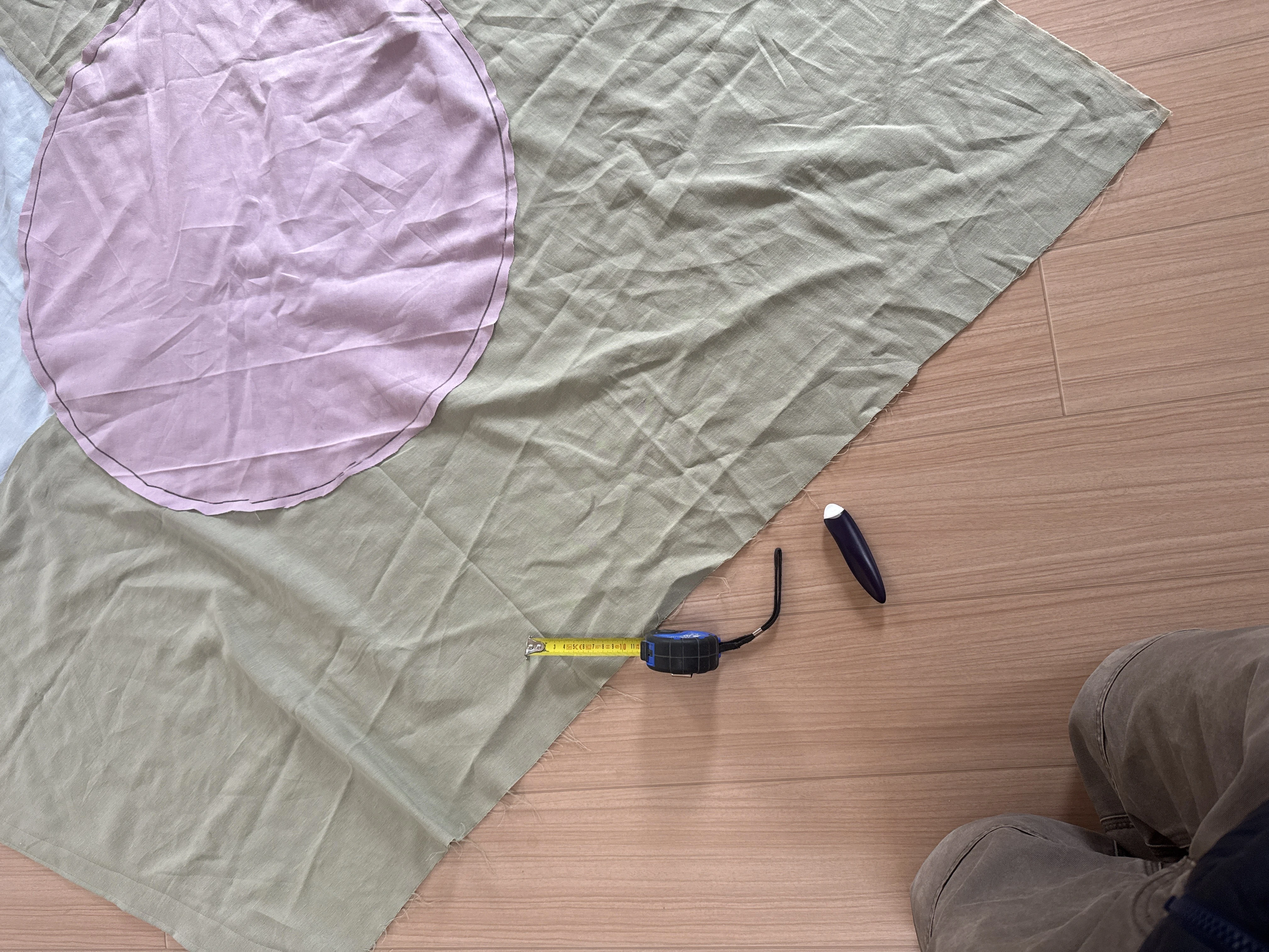 Sewing on a wood floor: sage green fabric with a pink circular piece, tape measure and chalk nearby